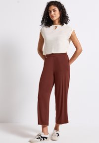 Crème top met een sluiting in de vorm van een sleutelgat, korte mouwen; roestkleurige, wijde broek; gecombineerd met witte sneakers met zwarte accenten.