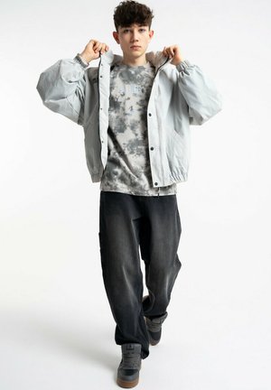 Młody mężczyzna poprawiający jasnoszary płaszcz, ubrany w szary t-shirt z efektem tie-dye, luźne ciemne spodnie i szare trampki, idzie do przodu na białym tle.