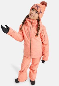Chaqueta de esquí de color coral brillante con capucha, combinada con pantalones a juego y guantes negros. El conjunto incluye un gorro de color naranja con un pom pom.