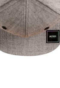 Grå caps med teksturert stoff, som har en flat skygge, og en svart lapp som viser "MSTRDS München"-logoen i hvitt og iriserende akenter.