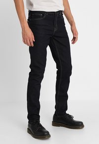 Tmavě modré džínové kalhoty slim fit, s pěti kapsami, kontrastním žlutým prošíváním a knoflíkovým zapínáním. Stylizované s černými šněrovacími botami.