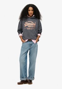 Sudadera gris con capucha y texto rosa, superpuesta sobre una camiseta rosa clara. Pantalones vaqueros sueltos de color azul claro con puños y zapatos de barco marrón oscuro.