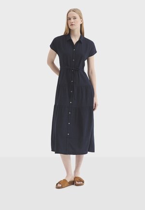 Kobieta ubrana w granatową sukienkę midi na guziki z paskiem w talii i krótkimi rękawami, w połączeniu z brązowymi sandałami wsuwanymi.