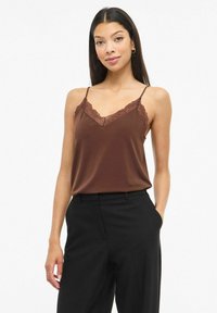 Brązowa satynowa koszulka na cienkich regulowanych ramiączkach z koronkową lamówką wzdłuż dekoltu, zestawiona z czarnymi, dopasowanymi spodniami.