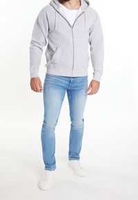 Sweat à capuche gris avec fermeture éclair et cordons, porté sur un t-shirt blanc. Associé à un jean skinny bleu clair et des baskets blanches.