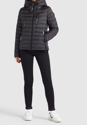 Žena ve černé prošívané bundě s kapucí, černých úzkých kalhotách a bílých teniskách, stojící na světlém pozadí.