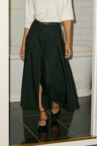 Jupe plissée noire à taille haute et fente latérale, assortie à un haut blanc et des chaussures noires. Tissu fluide, accentué par une ceinture.