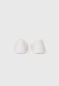 Une paire de baskets blanches vue de l'arrière sur un fond blanc uni, montrant les semelles texturées et le logo au talon.