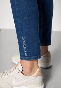 Mörkblå denimjeans med broderad logotyp "CALVIN KLEIN JEANS" på benet, i kombination med ljusa sneakers med bruna detaljer.