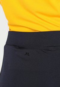 Tmavě modré sportovní šortky s hladkou texturou a elastickým pasem, s malým černým logem. Nad nimi je viditelná část žlutého topu.