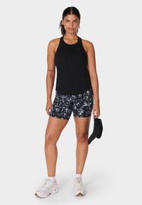 Vrouw in een zwarte mouwloze top en een patroonshort, die een zwarte pet vasthoudt, gekleed in witte sneakers, staand tegen een effen achtergrond.