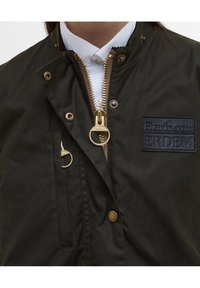 Donkergroene, gestructureerde jas met gouden ritssluiting en hardware, voorzien van een zwarte rubberen logopatch met de tekst "Barbour: ERDEM."