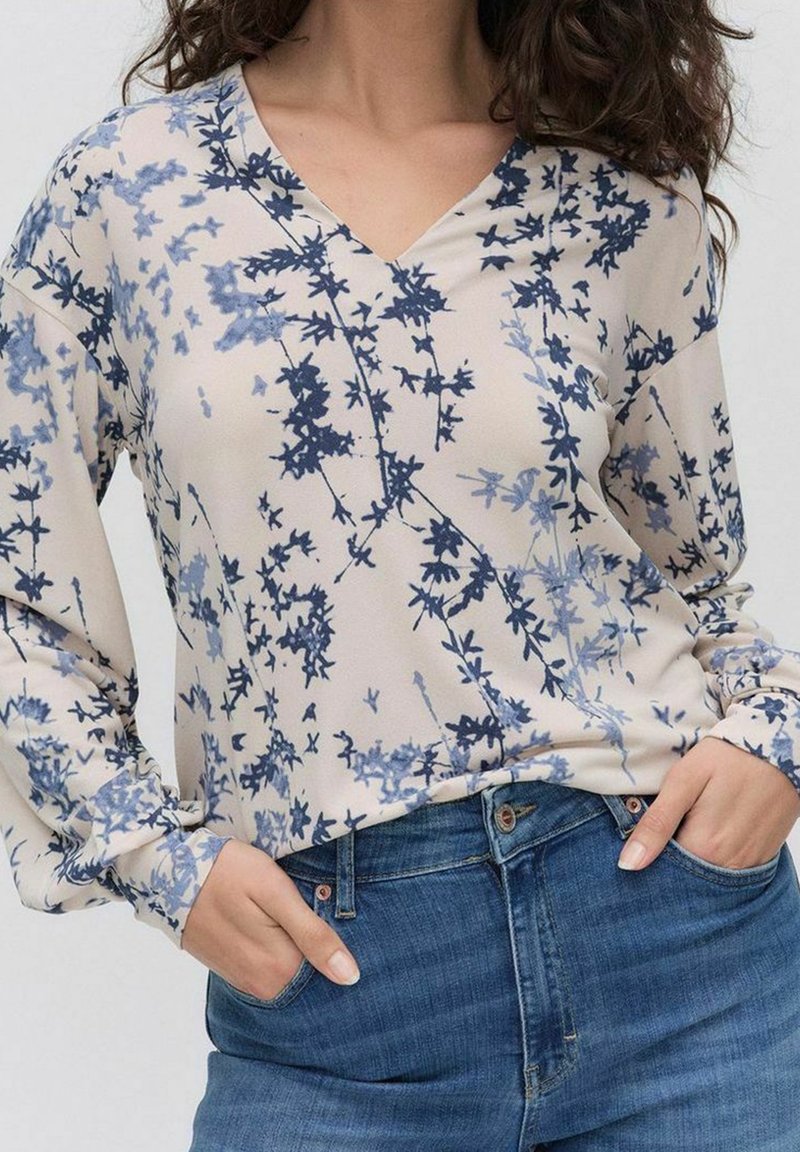 Blusenoberteil mit floralem Muster in Beige und blauen Akzenten, V-Ausschnitt und langen Ärmeln. Kombiniert mit klassischen blauen Jeans.