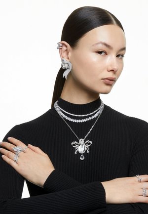 Schmuckset mit silberfarbenen Ketten, einem mit einem Edelstein verzierten Anhänger und kunstvollen Ohrklammern mit glitzernden Details, getragen von einem Model in einem schwarzen Oberteil.