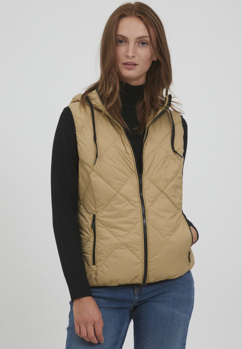Khakifarbenes, gestepptes Weste mit Kapuze, durchgehendem Reißverschluss und schwarzen Akzenten. Getragen über einem schwarzen Rollkragenpullover, kombiniert mit blauen Jeans. Glatte, leichte Textur.