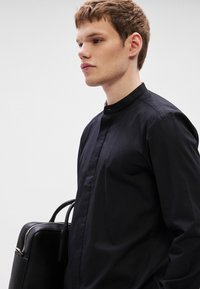 Fekete, galléros ing sima textilből, gombos záródással és letisztult kialakítással, fekete táskával, amelynek két fogófüle van.