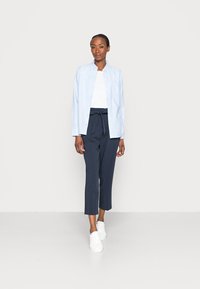 Camisa azul clara com botões, t-shirt branca por baixo e calças azul-marinho com a cintura amarrada, combinadas com sapatilhas brancas. Design simples e moderno.