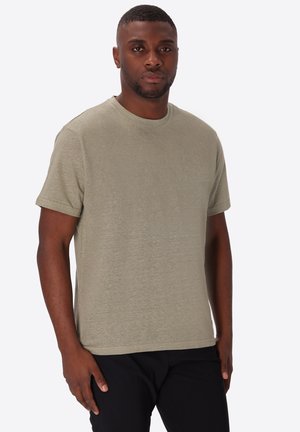 Uomo in piedi che indossa una maglietta beige tinta unita a maniche corte e pantaloni neri su sfondo bianco.