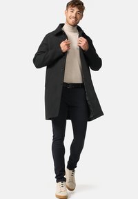 Ung mand smilende, iført beige højhalset trøje, sort frakke, sorte bukser og hvide sneakers, der justerer kraven på sin frakke, mens han går.