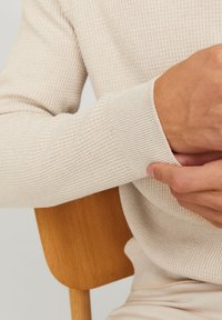 Ljusbeige långärmad ribbad tröja med strukturerat tyg; figursydda muddar och avslappnade ärmar; en hand placerad på handleden. Trästol i bakgrunden.