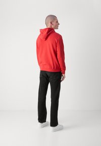 Felpa rossa con cappuccio, polsini e orlo a costine, abbinata a jeans neri e sneakers bianche. Modello di spalle rispetto alla fotocamera.