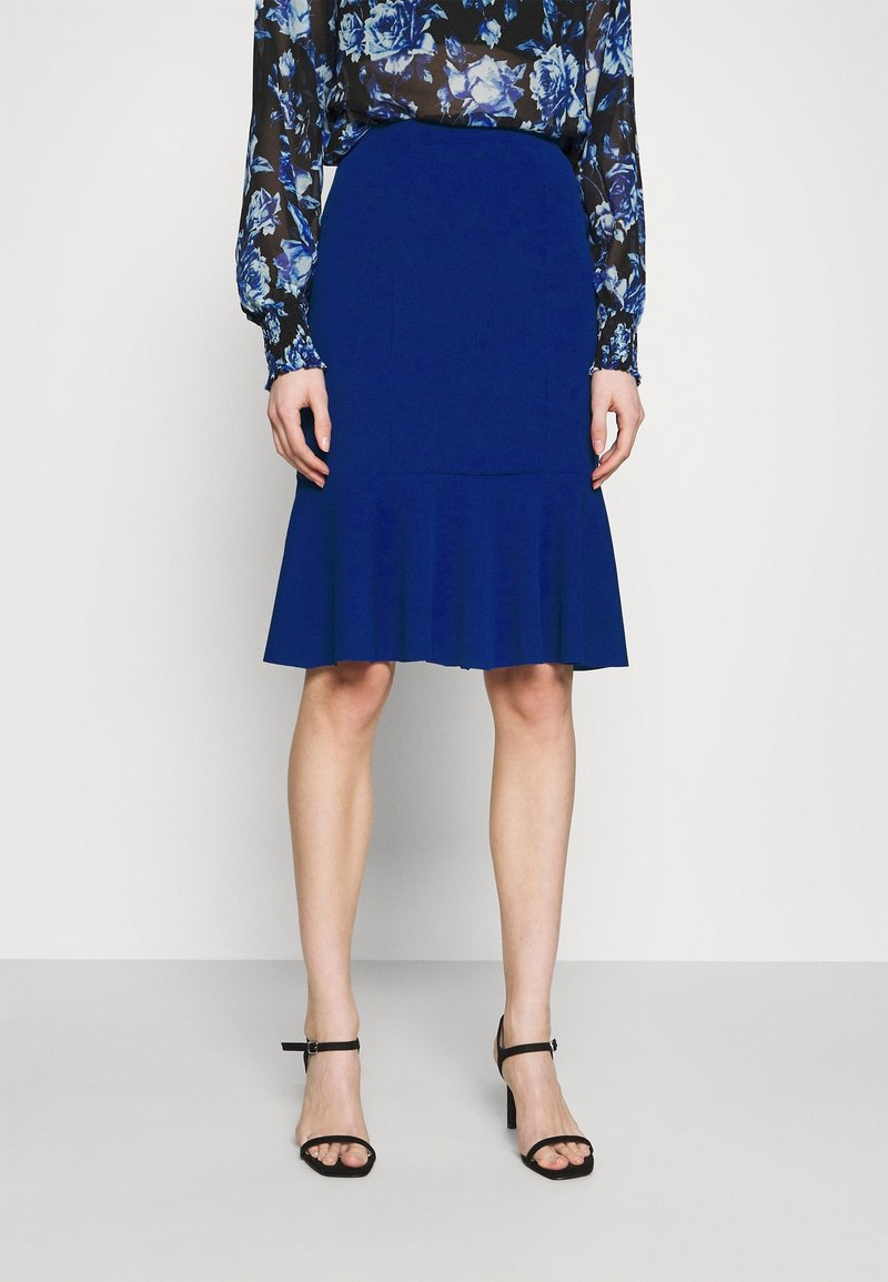 Jupe bleu ajustée avec un ourlet évasé, réalisée en tissu texturé, assortie à un chemisier noir à manches longues à motif floral et des talons noirs à brides.