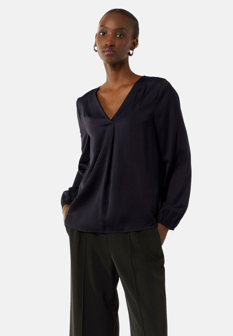 Granatowa bluzka z dekoltem w serek, z długimi rękawami i plisowanym designem. Wykonana z gładkiego materiału, z marszczonymi mankietami. W zestawie z ciemnymi spodniami.