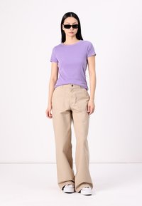 T-shirt viola a coste, vestibilità rilassata; pantaloni beige a gamba larga con tasche frontali; sneaker bianche; il modello indossa occhiali da sole neri.