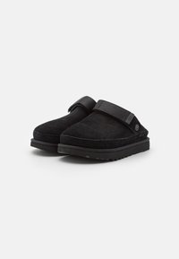 Ett par svarta slip-on mocka träskor med justerbara remmar och räfflade gummisulor mot en vit bakgrund.
