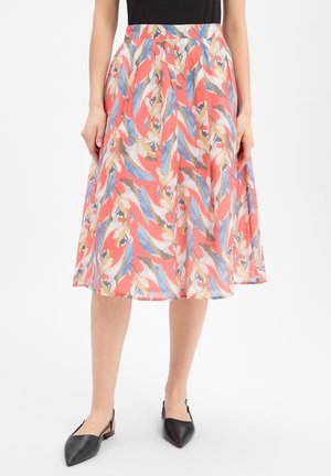 Vrouw draagt een koraalkleurige midi-rok met blauwe, beige en witte abstracte bladvormen, gecombineerd met zwarte puntige slingback schoenen.
