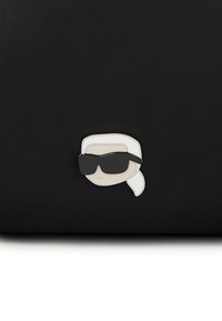 Svart stoffpose med en pin med et minimalistisk tegneserieansikt som har svarte solbriller og hvitt hår, som fremhever kontrast og designdetaljer.