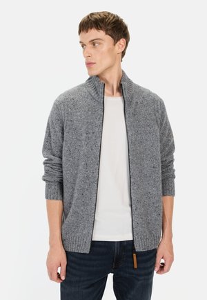 Graue gestrickte Strickjacke mit hohem Kragen, Reißverschluss, gerippten Bündchen und verstreuten Farbklecksen. Getragen über einem weißen Hemd und dunkler Jeans.