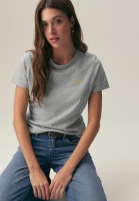 Sivo bombažno majico z okrogelim izrezom in kratkimi rokavi, okrašeno z majhnim rumenim vezenjem. Kombinirana z modrimi jeans hlačami in črnim pasom.