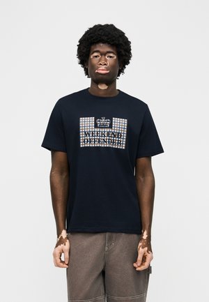 Νέος άνδρας με λευκά σημάδια δέρματος (βιτυλίγκο), φοράει μαύρο t-shirt με καρό λογότυπο κορώνας και το κείμενο "Weekend Offender", συνδυασμένο με χαλαρά καφέ παντελόνια.