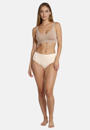 Donna in piedi a piedi nudi che indossa un reggiseno sportivo beige e biancheria intima crema a vita alta su uno sfondo bianco uniforme.
