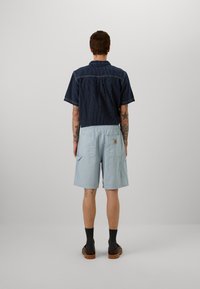 Pantaloni corti azzurro chiaro con finitura testurizzata e tasche laterali, abbinati a una camicia a righe navy a maniche corte, visti da dietro.