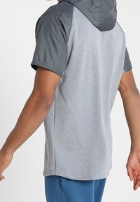 Bluza z kapturem z krótkim rękawem w jasnoszarym kolorze z ciemnoszarymi ramionami. Miękka tkanina, luźny krój i rozcięcie z tyłu zapewniające przewiewność.