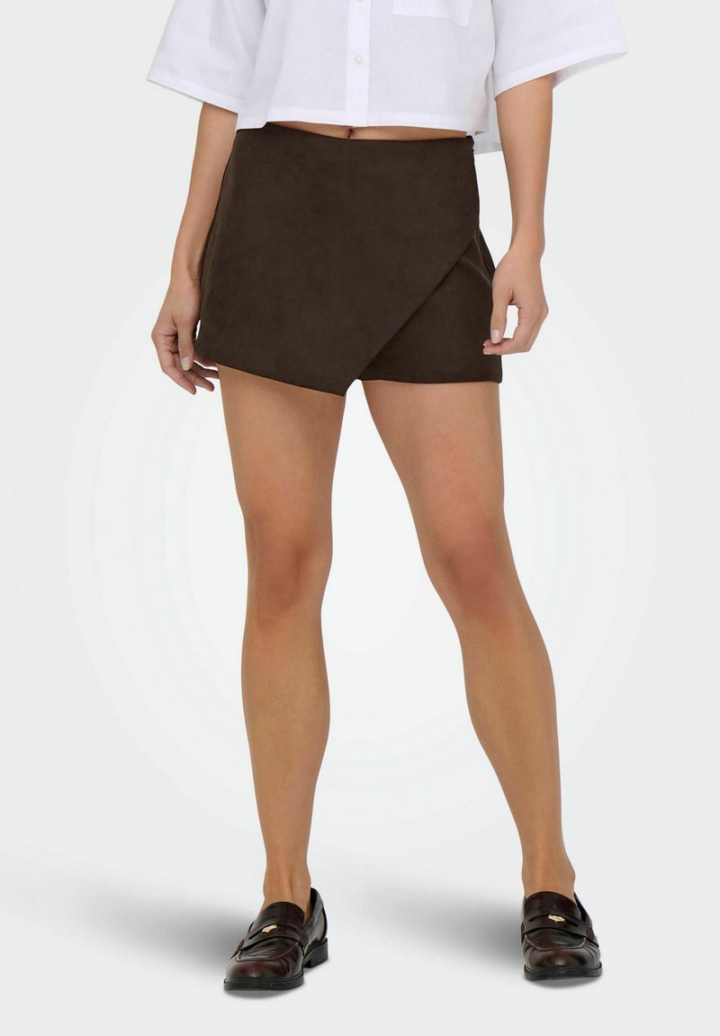 Kvinde iført en mørkebrun asymmetrisk miniskirt, hvid kortærmet skjorte og mørkebrun loafers, stående på en ensfarvet lys baggrund.