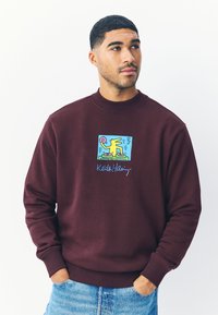 Vinröd sweatshirt med en färgglad Keith Haring-grafik av en DJ, med ribbade muddar och en lätt texturerad yta.