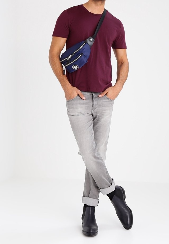 Homme portant un t-shirt bordeaux, un jean gris clair retroussé, des bottes noires et un sac bandoulière bleu avec des zips. Mains dans les poches, jambes croisées.