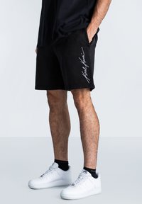 Melnie kokvilnas šorti ar brīvu piegriezumu, sānu kabatām un baltu paraksta logo uz kreisā gada. Apvienoti ar baltām sporta apaviem.
