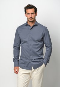 Camicia grigia a maniche lunghe con chiusura a bottoni, tessuto morbido e colletto a punta, indossata con pantaloni di colore chiaro.
