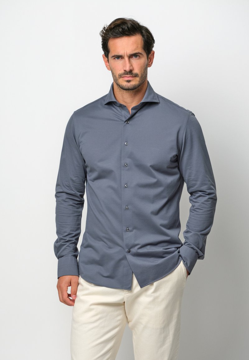 Camicia grigia a maniche lunghe con chiusura a bottoni, tessuto morbido e colletto a punta, indossata con pantaloni di colore chiaro.