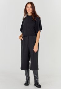 Juoda, trumpomis rankovėmis palaidinė su aukštu kaklu, derinama su juodomis plačiomis kelnėmis. Modelis dėvi juodus, iki kelių siekiančius batus. Lygus tekstūra.