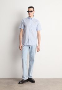 Homme portant des lunettes de soleil noires, une chemise bleu clair à manches courtes boutonnée, un jean bleu clair et des chaussures habillées noires, debout contre un mur blanc.