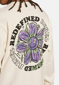 Sudadera de cuello redondo en beige claro con un gran gráfico de una flor púrpura y texto negro que dice "REDEFINIDO REBELDE" y "ROPA URBANA".