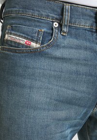 Jean en denim bleu avec une texture lisse, présentant des coutures orange, une poche avant et une étiquette de marque sur la poche arrière.