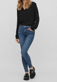 Maglietta nera a maniche lunghe, jeans skinny blu e mocassini neri. I jeans hanno una vita media e un leggero effetto sbiadito; i mocassini presentano dettagli in oro.