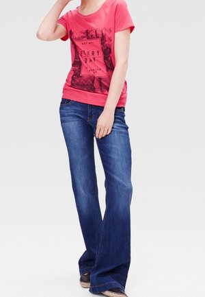 Kvinde iført en rød T-shirt med grafik og blå vide jeans, står med den ene hånd nær ansigtet og den anden hvilende ved siden.