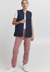 Quiltad marinblå ärmlös väst med hög halsringning. Bärs över en vit skjorta och matchas med ljusrosa sweatpants och vita sneakers.